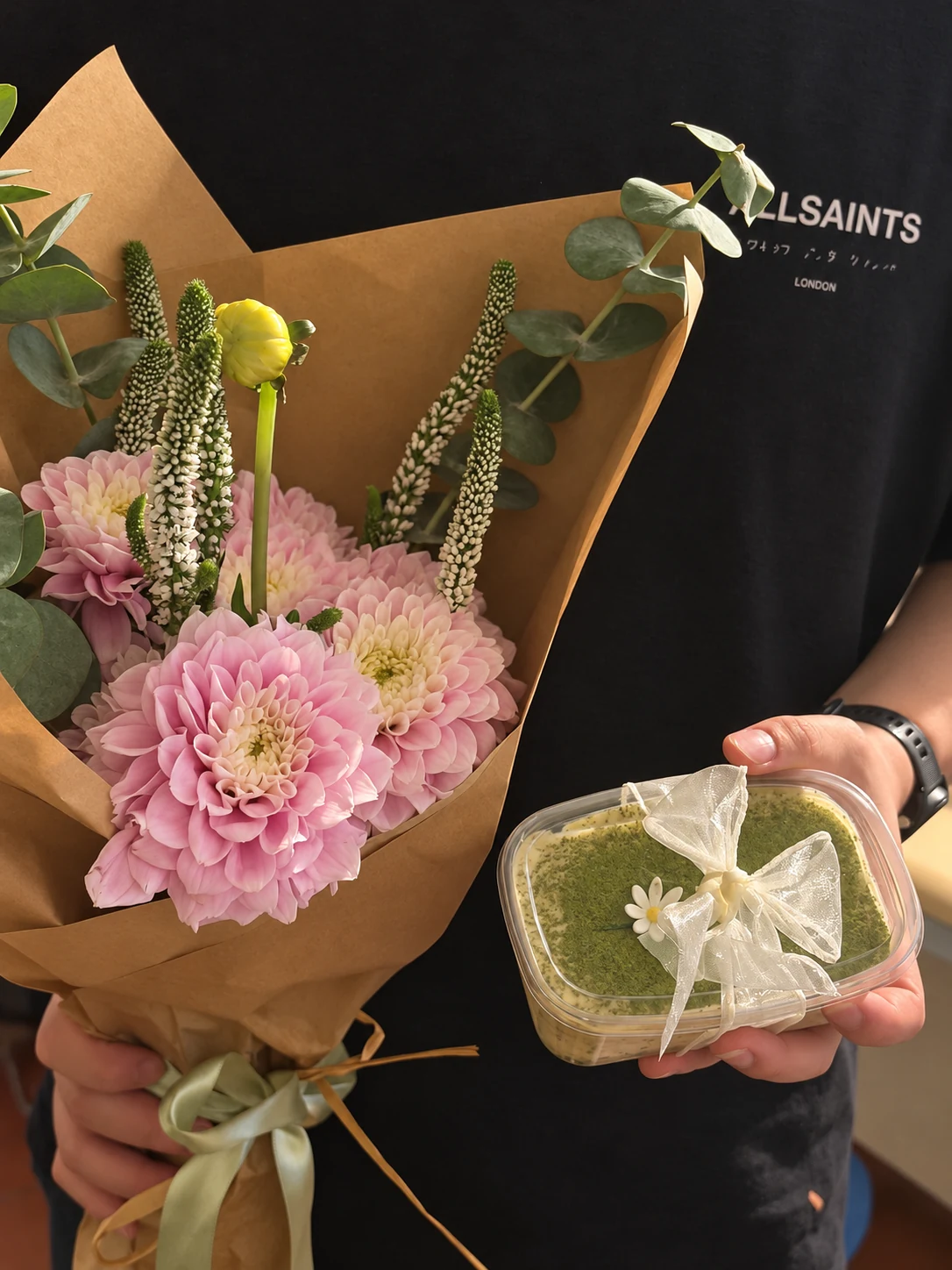 A ribbon tied around a matcha tiramisu cup, held beside pink flowers